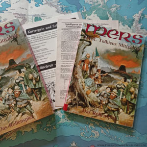 Auf dem Foto sind die MERS Box von Citadel, das Regelbuch, das Abenteuerbuch, Abenteuerpläne und eine farbige Mittelerde-Posterkarte abgelichtet.